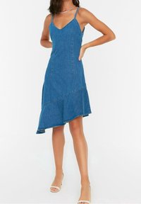 Denimklänning med V-ringning och tunna axelband, med en asymmetrisk nederkant. Färgen är medelblå med en strukturerad tyg.