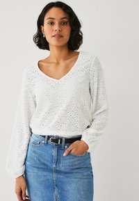 Blusa bianca in cotone a maniche lunghe con motivo a occhiello, scollo a V e polsini arricciati. Abbinata a una gonna blu in denim a vita alta e una cintura nera.