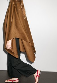 En brun ullponcho hänger över svarta byxor, med en löpande design och mjuk textur. Röda sandaler kompletterar outfiten.