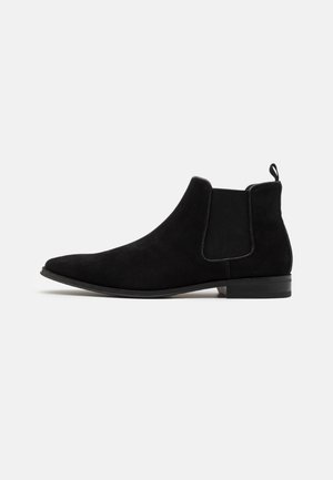 Bottines en suède noir avec des panneaux élastiques sur les côtés, un bout pointu et une languette à l'arrière. Semelle plate et design minimaliste.
