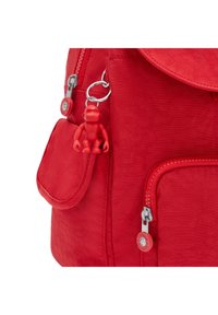 Mochila roja con textura suave, que presenta un cierre de solapa, bolsillo frontal, herrajes de cremallera plateados y una pequeña figura de juguete roja adjunta.