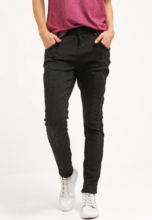 Persoon die strakke zwarte distressed jeans draagt, een roze-paarse top en witte veterschoenen, staand met handen in de zakken.