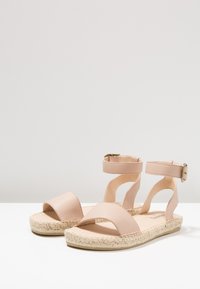 Beige flat sandals with ankle straps and buckles, open toes, and woven espadrille-style soles, displayed on a white surface.