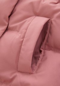 Detail einer rosa gepolsterten Jacke mit einer glatten, matten Textur und einer Seitentasche mit sauberem Nähen. Der Stoff wirkt leicht und dennoch warm.