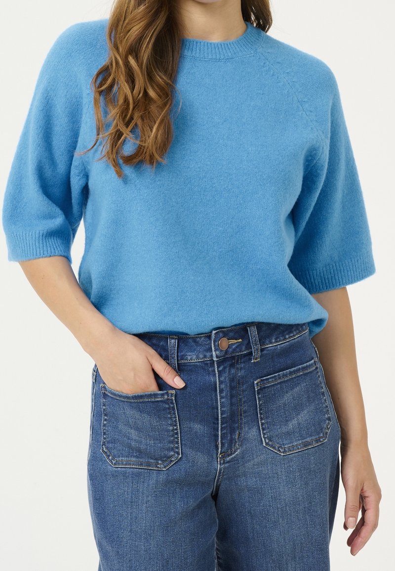 Femme portant un pull en maille bleu à manches courtes rentré dans un jean taille haute bleu avec des poches plaquées frontales, main dans la poche gauche.