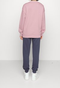 Persona con sudadera rosa, pantalones deportivos azul marino y zapatillas blancas de pie de espaldas sobre un fondo blanco liso.