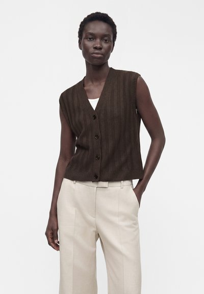 Femme portant un gilet sans manches côtelé marron foncé boutonné et un pantalon beige taille haute sur un fond uni.