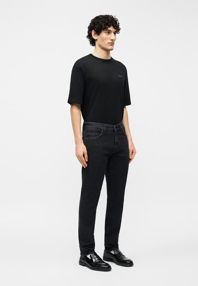 Νεαρός άνδρας με σγουρά μαλλιά που φοράει απλό μαύρο μπλουζάκι, σκούρα στενά τζιν και μαύρα δερμάτινα μοκασίνια, στέκεται μπροστά σε μια απλή φόντο.