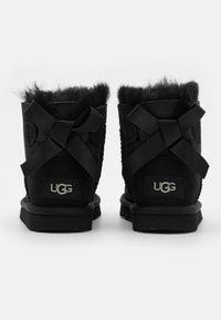 Bottines en suède noir avec doublure en laine pelucheuse et nœuds en satin noir à l'arrière. Présente un logo de marque visible sur le talon.