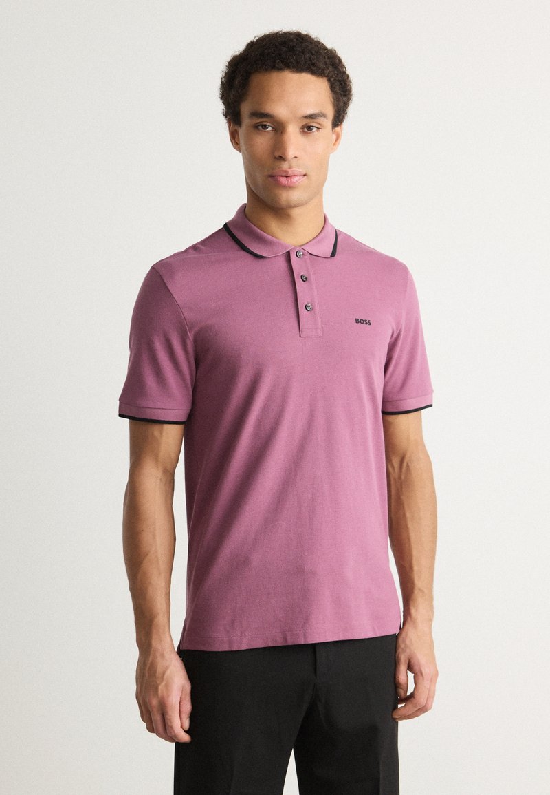Polo majica s kratkimi rokavi v barvi mauve z ovratnikom in manšetami obrobljenimi v črni barvi, ki vključuje tri gumbke na zapenjanju in majhno logotip na prsih.
