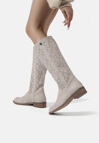 Stivali in suede beige alti fino al ginocchio con un design a fioritura intagliata al laser, tacco piatto e suola testurizzata, caratterizzati da un dettaglio decorativo a bottone sul lato.