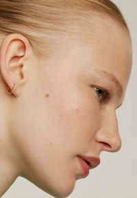 Pendiente de aro de oro con pequeñas piedras preciosas. Textura de piel suave, color de labios rosa suave y cabello rubio claro, mostrado en perfil lateral.