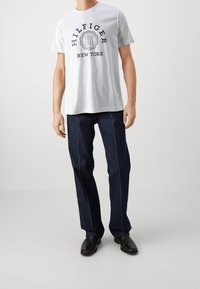 Camiseta blanca de algodón con gráfico negro 'HILFIGER NEW YORK', combinada con pantalones de mezclilla azul oscuro con costuras azul claro y zapatos negros.