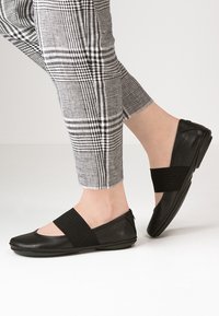 Zapatos planos negros con correas elásticas, con una textura similar al cuero suave, punta redondeada y suela flexible, combinados con pantalones a cuadros.