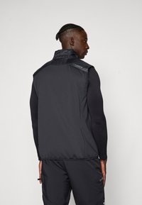 Melna veste ar paklājotu plecu un apkakli. Iezīmējas ar gludu materiālu un pieguļošu dizainu, kas sasniedz jostasvietu. Logotips ir redzams uz pleca.