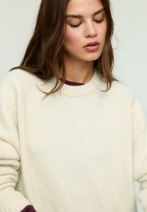 Cream-colored knit sweater with a round neckline, ribbed cuffs, and soft texture. The fabric appears warm and cozy, with a slightly relaxed fit.