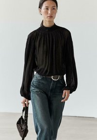 Sort bluse med lange ærmer og h�øj smocket hals, kombineret med højttaljede blå jeans og en mørk håndtaske. Glat tekstur, afslappet pasform.