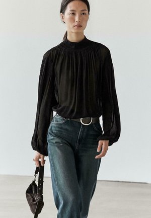 Czarna bluzka z długim rękawem i wysokim marszczonym kołnierzem, zestawiona z wysokim stanem niebieskich jeansów i ciemną torebką. Gładka tekstura, luźny krój.
