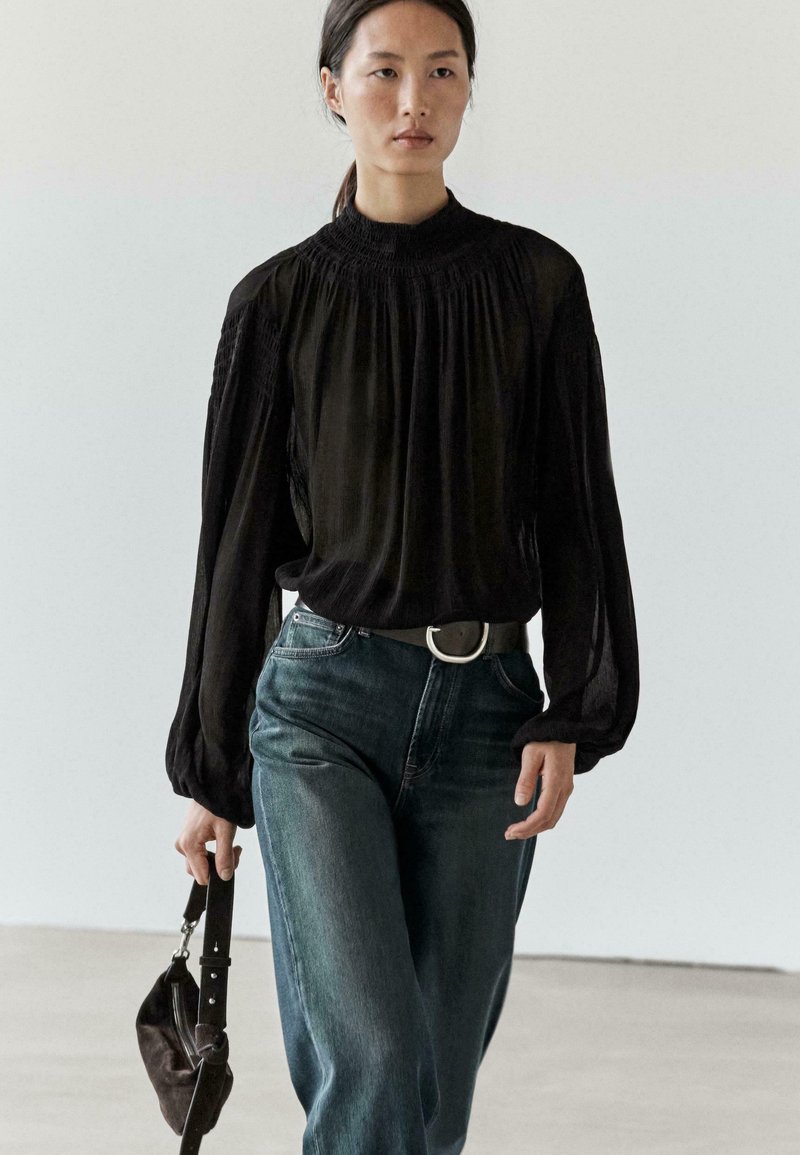 Sort bluse med lange ærmer og høj smocket hals, kombineret med højttaljede blå jeans og en mørk håndtaske. Glat tekstur, afslappet pasform.