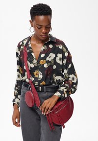 Femme portant un blouse florale noire et un pantalon gris, accessoirisée avec un sac bandoulière rouge et un petit étui rond, regardant vers le bas.
