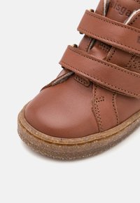 Chaussure en cuir marron avec deux sangles Velcro, bout arrondi et semelle en caoutchouc texturée. Des détails cousus et un intérieur doux sont visibles.