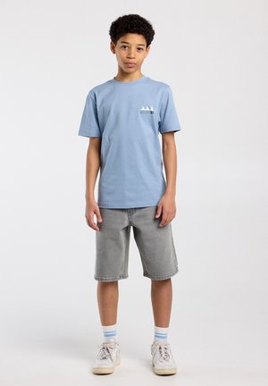 Jongen staand, gekleed in een lichtblauw T-shirt, grijze korte broek, witte sneakers en witte sokken met blauwe strepen, tegen een effen achtergrond.