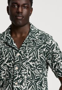 Camicetta a maniche corte realizzata in un tessuto morbido, con un motivo a foglie verde e bianco, chiusura con bottoni e un design del colletto rilassato.