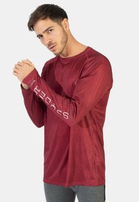 Pitkähihainen urheilusade burgundin värissä, jossa on teksturoitu pinta, tasaiset saumat ja valkoinen logo yksityiskohtina hihassa.