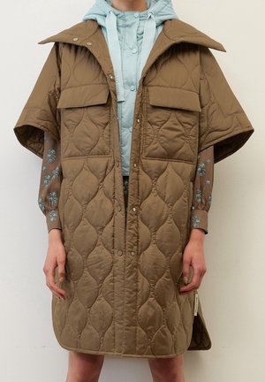 Persoon draagt een bruine gewatteerde mouwloze jas over een blauwe capuchontrui en een bruin bloes met lange mouwen, staand tegen een witte achtergrond.