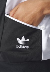 Svart och vit Adidas dragkedjest Pocket i texturerad tyg; har tre-stripes logotyp i vitt, accentuerad med en slät dragkedjestängning.