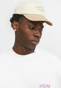 Beige pet met een gebogen klep en geborduurde tekst "ORIGINALS ATELIER." Het model draagt een wit t-shirt met een roze logo. Close-up weergave.