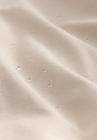 Beiger Stoff mit kleinen Wassertröpfchen, die auf der Oberfläche perlen und die wasserabweisende Textur sowie leichte Falten hervorheben.