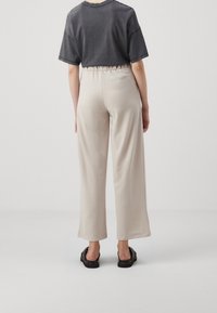 Person med beige vida byxor, svarta sandaler och en mörkgrå kortärmad skjorta, sedd bakifrån mot en neutral bakgrund.