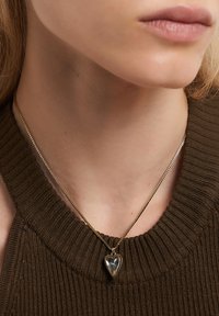 Guldfärgad halsband med ett hjärtformat hängsmycke som har en genomskinlig mitt, buren på en ribbad, mörkbrun tröja.