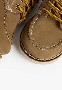 Braune Wildleder-Stiefeletten mit gelben Schnürsenkeln, kontrastierenden weißen Nähten und einer robusten Gummisohle. Rundliche Zehenpartie und klassisches Mokassin-Design.