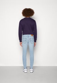 Sudadera morada, jeans ajustados azul claro, tela texturizada, vista trasera. Parche de cuero marrón notable en la cinturilla, zapatillas blancas.