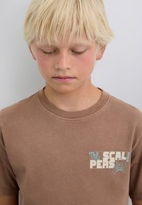 Niño rubio con los ojos cerrados que lleva una camiseta marrón con el texto "Los Scalpers" y un gráfico de calavera con huesos cruzados.