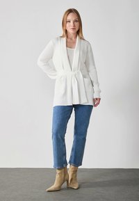 White, ribbed cardigan with a tied waist, two front pockets, and open front. Paired with blue jeans and beige heeled ankle boots.