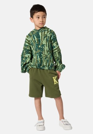 Sweat à capuche vert à motifs avec design de feuilles, associé à un short vert olive avec un accent jaune "K". Chaussures blanches.