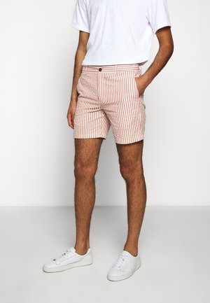 Mann trägt weiße Sneakers, pink-weiß vertikal gestreifte Shorts und ein schlichtes weißes Kurzarmshirt, steht mit einer Hand in der Tasche.