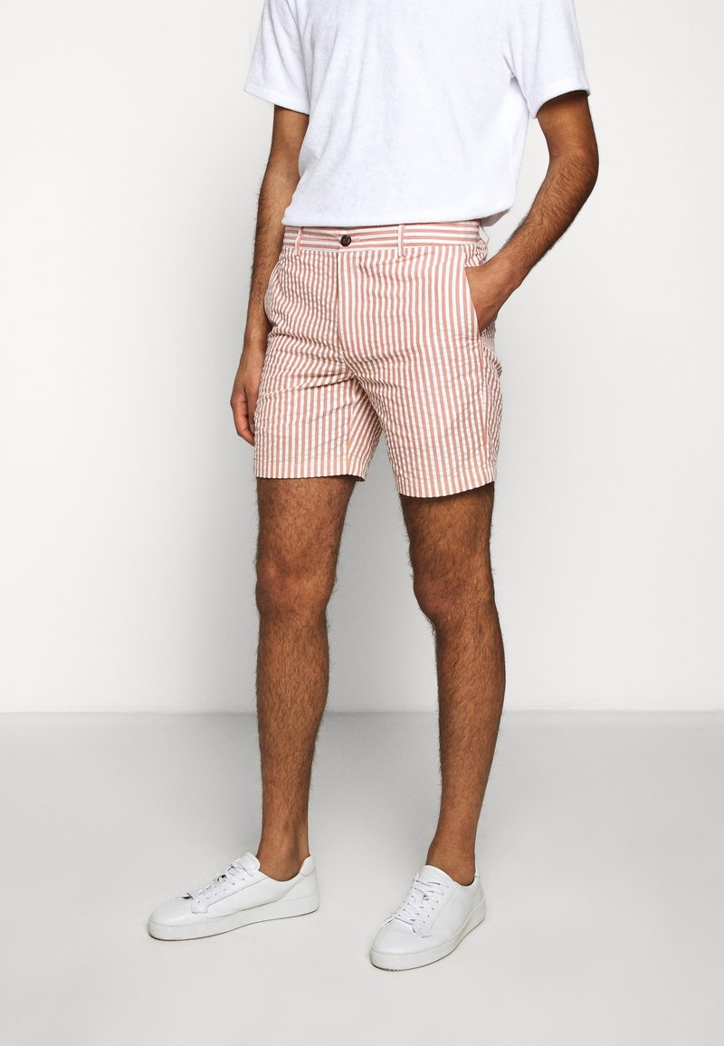 Homme portant des baskets blanches, un short rayé vertical rose et blanc, et un t-shirt blanc uni à manches courtes, debout avec une main dans la poche.