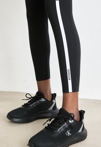 Zwarte legging met witte zijstrepen, met "Calvin Klein" branding; gecombineerd met zwarte mesh sport schoenen en gestructureerde zolen.