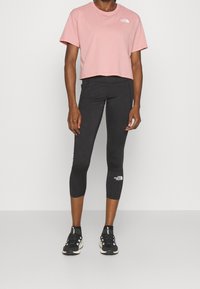 Gekruiste roze T-shirt met een wit logo, gecombineerd met zwarte leggings met een wit logo, en zwarte sportschoenen met witte accenten.