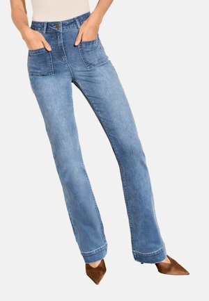 Kvinde iført lyseblå lige jeans med store forlommer og brune spidse hæle, hænderne placeret i lommerne.