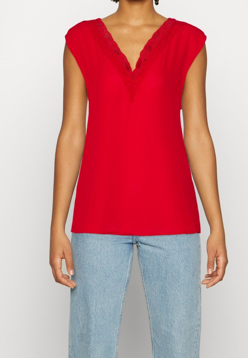 Femme portant un haut rouge sans manches avec un col en V en dentelle, associé à un jean bleu clair, debout devant un fond clair uni.