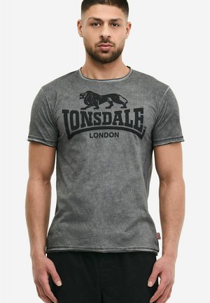 Mann trägt ein verblasstes graues Lonsdale London T-Shirt mit schwarzem Löwen-Logo, steht mit entspannten Armen an den Seiten.