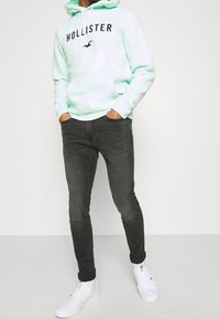 Mintgrön huvtröja med justerbara snören och framficka, kombinerad med mörkgrå slim-fit jeans och vita sneakers. Svart logotyp på huvtröjan.