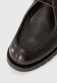Close-up van een donkerbruin leren schoen met gedetailleerde stiksels en een dikke zwarte rubberen zool op een witte achtergrond.