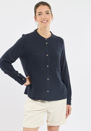 Femme souriante portant une chemise bleu marine à manches longues avec deux poches à l'avant et un short beige retroussé, se tenant debout la main sur la hanche.
