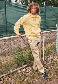 Yellow hoodie with a front pocket and logo, paired with beige pants featuring a logo patch, and black sneakers with pink accents.